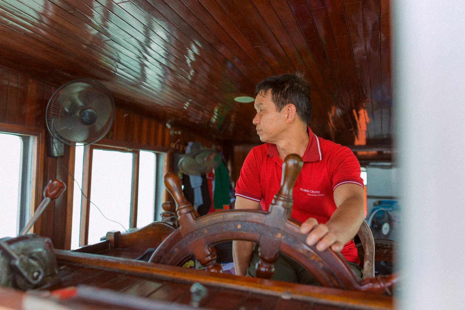 Man in red shirt steering a traditional wooden boat, showcasing maritime navigation skills.