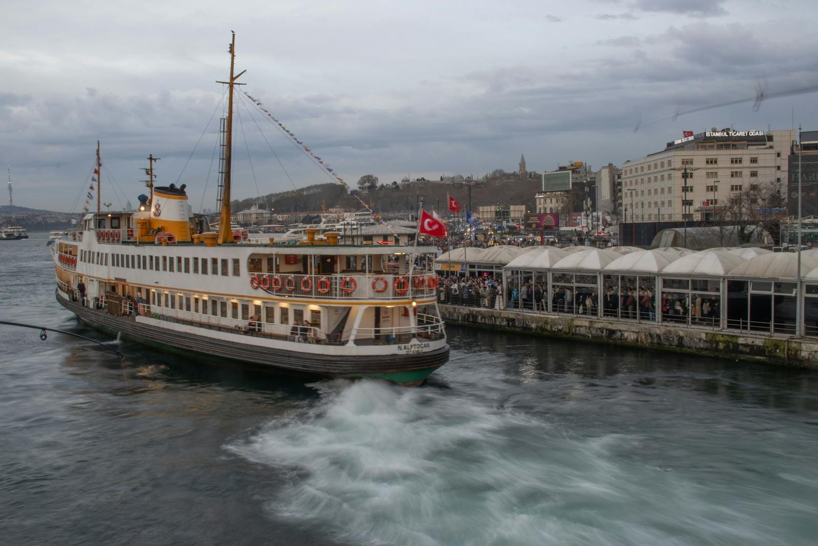 An iconic ferry approaches the bustling Eminönü port in Istanbul, Turkey, under a cloudy sky.